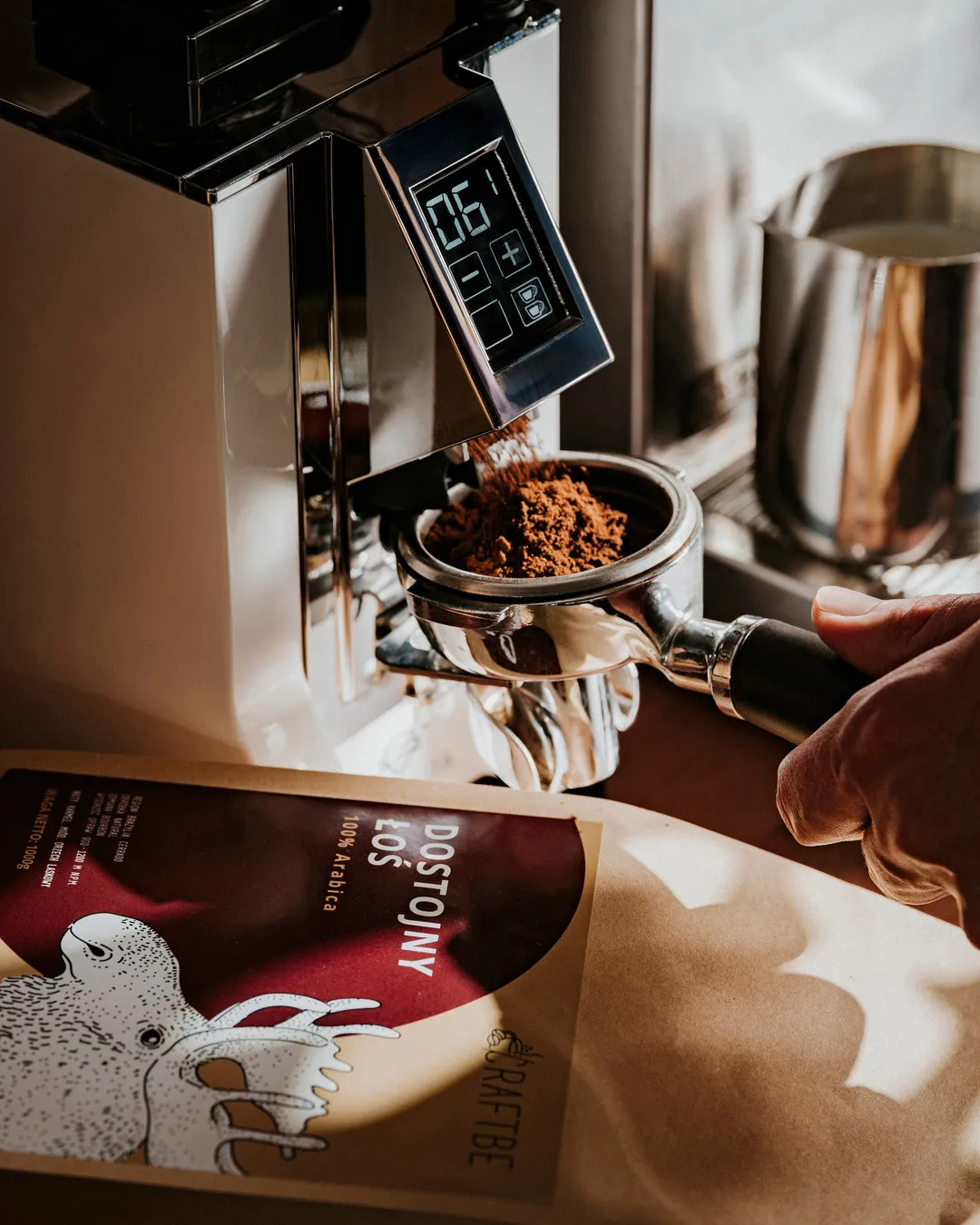 Młynek sypie świeżo zmieloną kawę do kolby ekspresu; obok Dostojny Łoś 1 kg