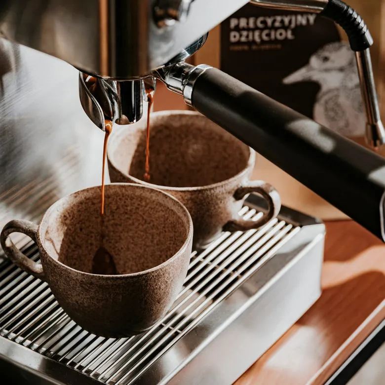 Espresso wypływa z kolby ekspresu do dwóch kamionkowych filiżanek, w tle paczka Precyzyjnego Dzięcioła
